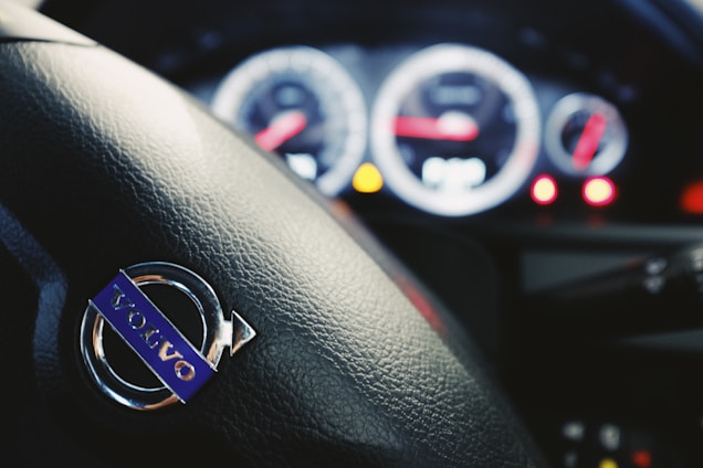 The close-up of a car steering wheel featuring the Volvo logo with dashboard dials and indicators blurred in the background.