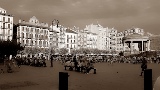 A bustling city square featuring historic European architecture. Numerous people are gathered, some sitting on benches while others are walking around the open space. A prominent gazebo or bandstand can be seen to the right, adding charm to the scene. The image has a sepia tone, giving it a vintage feel. Streetlamps and trees line the background alongside multi-story buildings with decorative facades.