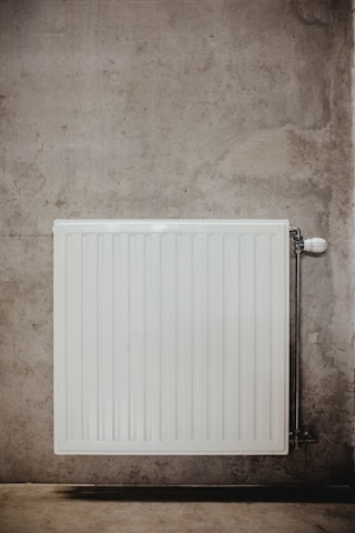 A white, rectangular radiator is mounted on a concrete wall, featuring vertical ridges and a valve on the right side. The concrete surface is textured with subtle variations in color and roughness.