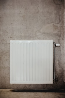 A white, rectangular radiator is mounted on a concrete wall, featuring vertical ridges and a valve on the right side. The concrete surface is textured with subtle variations in color and roughness.
