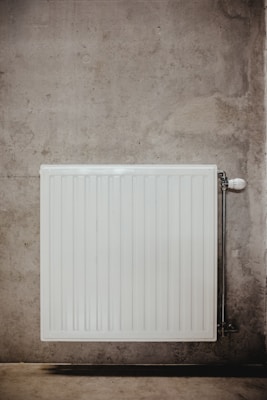 A white, rectangular radiator is mounted on a concrete wall, featuring vertical ridges and a valve on the right side. The concrete surface is textured with subtle variations in color and roughness.