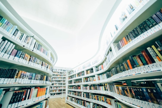 A modern library interior with curved, white bookshelves filled with a vast array of colorful books. The design creates a feeling of openness and spaciousness, accentuated by the softly lit ceiling. The floor is made of light wood, adding warmth to the environment.