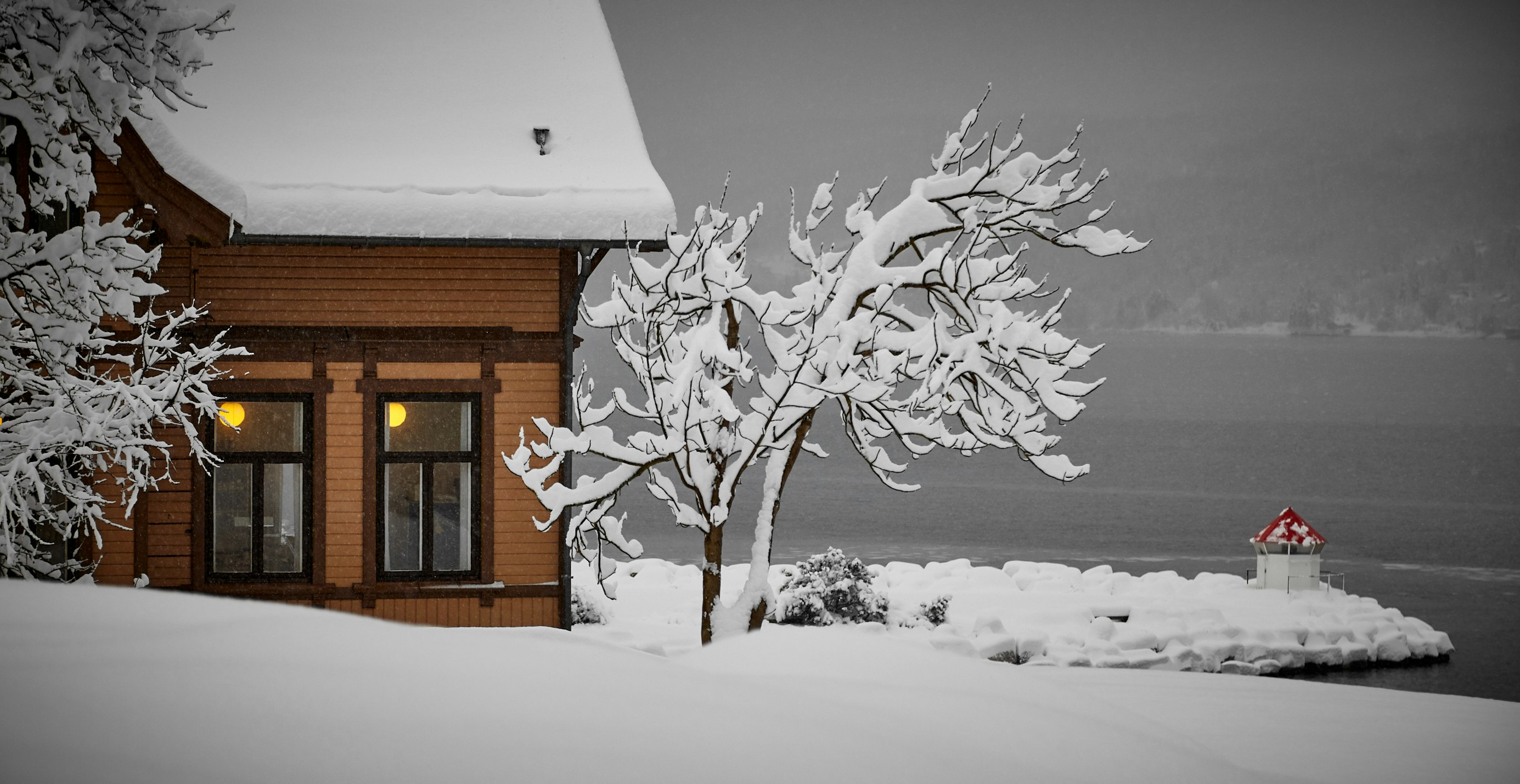 snow covered house on gray lake with lights on in the window