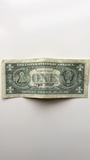 A worn United States one-dollar bill is centered on a plain background. The bill bears the typical symbols and inscriptions found on U.S. currency, including 'The United States of America' and 'In God We Trust'. However, the text 'F*CK TRUMP' is handwritten in black ink across the word 'ONE'.