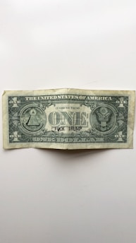 A worn United States one-dollar bill is centered on a plain background. The bill bears the typical symbols and inscriptions found on U.S. currency, including 'The United States of America' and 'In God We Trust'. However, the text 'F*CK TRUMP' is handwritten in black ink across the word 'ONE'.