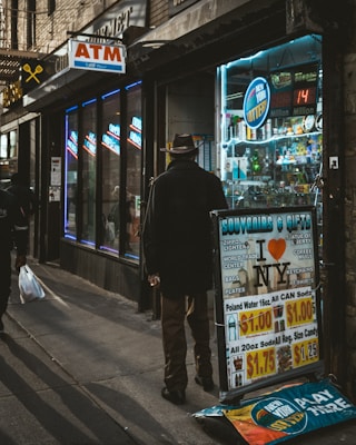 A dimly lit street scene featuring a man wearing a hat and coat standing near a storefront with neon signs. The store advertises souvenirs and gifts, with posters for NYC-themed items like t-shirts and keychains. An ATM sign and New York lottery signage are visible, along with various items displayed inside the store.