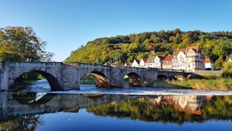 A stone bridge with multiple arches spans across a calm river, with reflections of the bridge and nearby buildings visible in the water. A row of charming, traditional houses with sloped roofs sits adjacent to a densely wooded hillside, where the foliage displays various shades of green. People can be seen leisurely walking across the bridge.