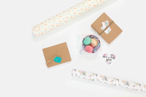 A light and airy arrangement of gift wrapping materials. Two small packages wrapped in brown paper are tied with twine. Nearby, a small nest filled with colorful macarons is surrounded by pastel paper shavings. Rolls of decorative paper with floral and polka dot patterns are positioned diagonally. A heart-shaped card with floral designs and the text 'I need you' adds a touch of sentiment.