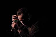 A person kneeling in front of a microphone in low lighting, casting shadows on their face. The mood is contemplative and serious.