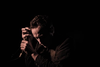 A person kneeling in front of a microphone in low lighting, casting shadows on their face. The mood is contemplative and serious.