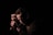 A person kneeling in front of a microphone in low lighting, casting shadows on their face. The mood is contemplative and serious.