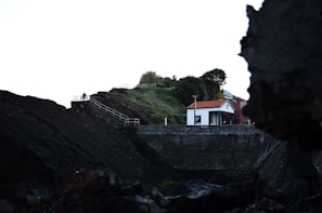 A small white house with a red roof is situated on a cliff surrounded by dark rocky terrain and green vegetation. A staircase with railings leads up the cliff, and a person is visible sitting near the top. The scene is framed by large rocks in the foreground.