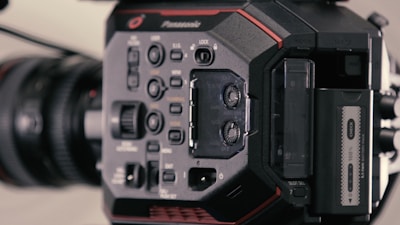 A close-up view of a professional video camera, prominently displaying its control panel with various buttons and dials. The lens is visible to the left, and there are slots for memory cards and other audio-visual input options. The design is sleek with a mix of black and red colors.