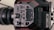 A close-up view of a professional video camera, prominently displaying its control panel with various buttons and dials. The lens is visible to the left, and there are slots for memory cards and other audio-visual input options. The design is sleek with a mix of black and red colors.