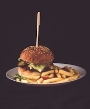 A hamburger with lettuce, pickles, and cheese is positioned at the center of a white plate, accompanied by a serving of golden-brown French fries. A skewer is inserted through the hamburger, which is topped with a sesame seed bun. The background is dark, emphasizing the vibrant colors of the food.