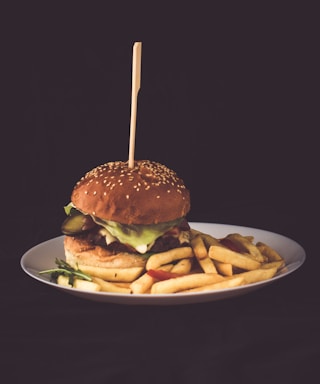 A hamburger with lettuce, pickles, and cheese is positioned at the center of a white plate, accompanied by a serving of golden-brown French fries. A skewer is inserted through the hamburger, which is topped with a sesame seed bun. The background is dark, emphasizing the vibrant colors of the food.