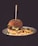 A hamburger with lettuce, pickles, and cheese is positioned at the center of a white plate, accompanied by a serving of golden-brown French fries. A skewer is inserted through the hamburger, which is topped with a sesame seed bun. The background is dark, emphasizing the vibrant colors of the food.