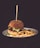 A hamburger with lettuce, pickles, and cheese is positioned at the center of a white plate, accompanied by a serving of golden-brown French fries. A skewer is inserted through the hamburger, which is topped with a sesame seed bun. The background is dark, emphasizing the vibrant colors of the food.
