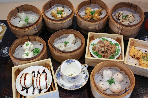 A variety of dim sum dishes presented in bamboo steamers and wooden trays. Dishes include dumplings, buns, and other bite-sized portions, each garnished with herbs. A floral-patterned tea cup with tea is placed prominently in the center among the food.