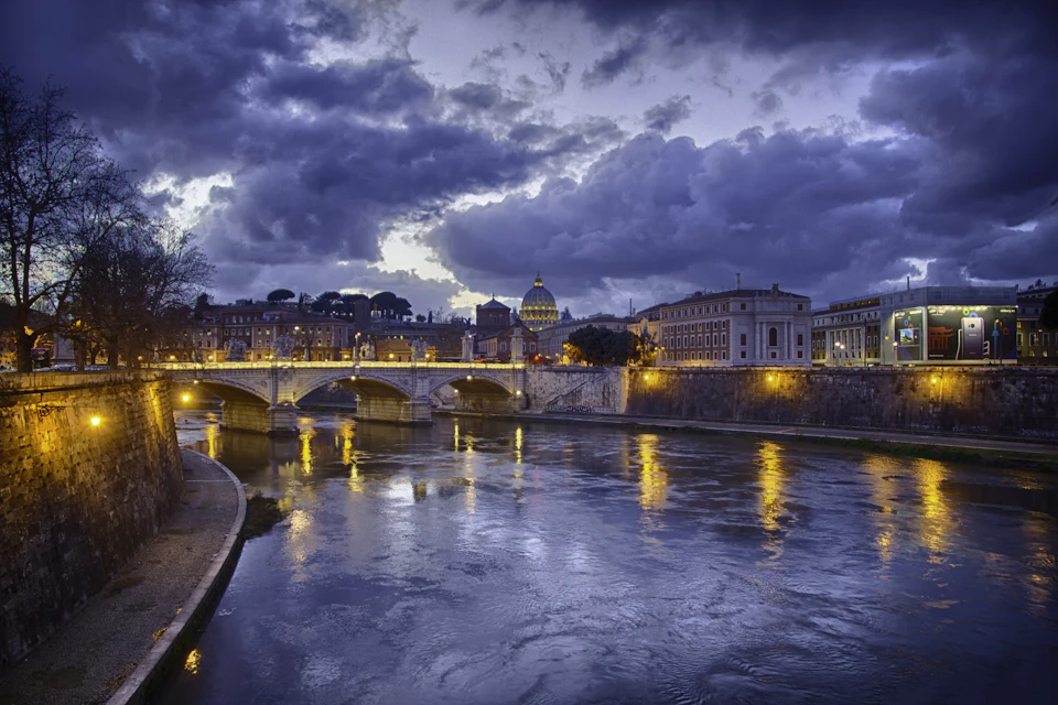 The Tiber at dusk under heavy grey cloud, lamps lit along the embankment, a stone bridge with St Peter's dome beyond it and bare trees on the left bank