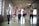 Six women are exercising in a spacious, light-filled gym with exposed brick walls and large windows. They are standing in a row, each holding a pink dumbbell in one hand, and appear to be engaged in a fitness routine. The women are dressed in athletic wear, including sports bras and leggings, in a variety of colors. The room has a high, wooden-beamed ceiling and a polished concrete floor.