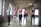 Six women are exercising in a spacious, light-filled gym with exposed brick walls and large windows. They are standing in a row, each holding a pink dumbbell in one hand, and appear to be engaged in a fitness routine. The women are dressed in athletic wear, including sports bras and leggings, in a variety of colors. The room has a high, wooden-beamed ceiling and a polished concrete floor.