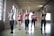 Six women are exercising in a spacious, light-filled gym with exposed brick walls and large windows. They are standing in a row, each holding a pink dumbbell in one hand, and appear to be engaged in a fitness routine. The women are dressed in athletic wear, including sports bras and leggings, in a variety of colors. The room has a high, wooden-beamed ceiling and a polished concrete floor.
