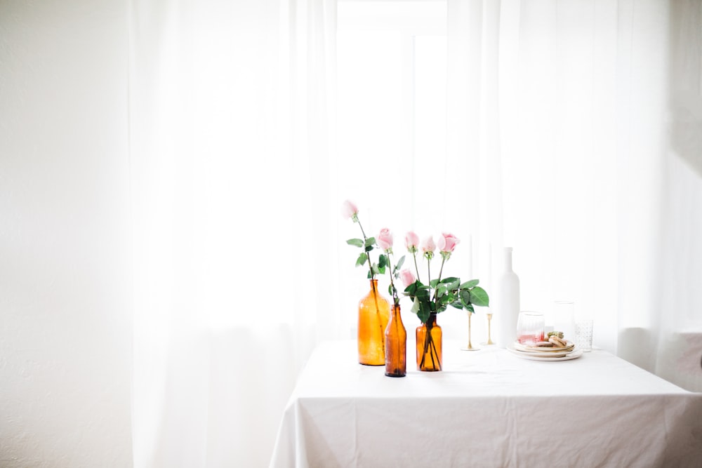 Drei Braunglas-Blumenvasen mit rosa Blumen auf weißem Tisch in der Nähe des Fenstervorhangs