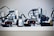 Several LEGO Mindstorms robots are lined up on a shelf with visible wires and sensor modules. Each robot has wheels and complex assemblies made from LEGO pieces, including sensors and connectors.