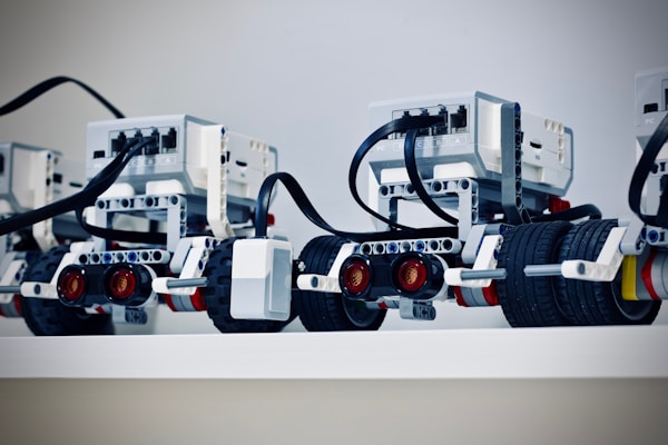 Several LEGO Mindstorms robots are lined up on a shelf with visible wires and sensor modules. Each robot has wheels and complex assemblies made from LEGO pieces, including sensors and connectors.
