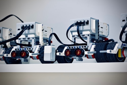 Several LEGO Mindstorms robots are lined up on a shelf with visible wires and sensor modules. Each robot has wheels and complex assemblies made from LEGO pieces, including sensors and connectors.