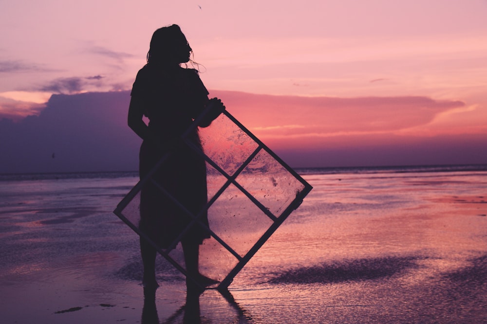 silhouette of woman holding windowpane on shore during golden hour