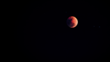 A dark sky features a prominent celestial body that appears to be a red or blood moon, partially obscured by shadow. The surrounding space is vast and mostly black with faint stars visible around the moon.