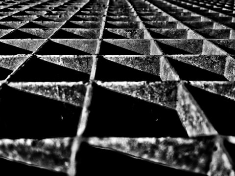 A geometric pattern of metallic grid structures with triangular openings, featuring a high-contrast black and white color scheme and dramatic shadows.