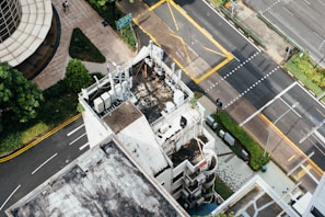 Aerial view of a city intersection with roads, sidewalks, buildings, and greenery. Several air conditioning units and other equipment can be seen on the rooftop of a building in the foreground. The streets are marked with white and yellow lines, and there are crosswalks and traffic signs visible.