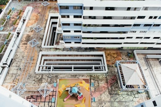 An aerial view of a residential building complex, showcasing multiple floors of a modern white and blue multi-story building. Below, there is a geometric, colorful playground area with playground equipment including slides and climbing structures. The surrounding concrete courtyard has multiple covered walkways and geometric metal structures, possibly shading or seating areas. The ground appears worn and weathered with visible cracks and patterns.