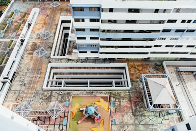 An aerial view of a residential building complex, showcasing multiple floors of a modern white and blue multi-story building. Below, there is a geometric, colorful playground area with playground equipment including slides and climbing structures. The surrounding concrete courtyard has multiple covered walkways and geometric metal structures, possibly shading or seating areas. The ground appears worn and weathered with visible cracks and patterns.