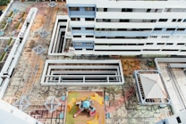 An aerial view of a residential building complex, showcasing multiple floors of a modern white and blue multi-story building. Below, there is a geometric, colorful playground area with playground equipment including slides and climbing structures. The surrounding concrete courtyard has multiple covered walkways and geometric metal structures, possibly shading or seating areas. The ground appears worn and weathered with visible cracks and patterns.