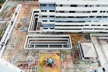 An aerial view of a residential building complex, showcasing multiple floors of a modern white and blue multi-story building. Below, there is a geometric, colorful playground area with playground equipment including slides and climbing structures. The surrounding concrete courtyard has multiple covered walkways and geometric metal structures, possibly shading or seating areas. The ground appears worn and weathered with visible cracks and patterns.