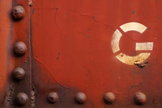 A rusty metal surface with visible rivets and a faded white circular emblem featuring segments. The surface shows signs of corrosion and aging.