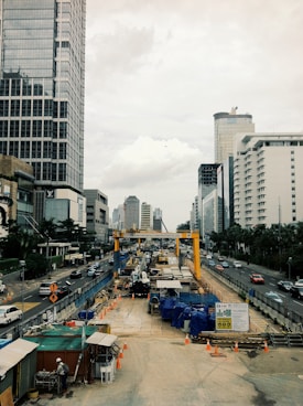 A busy city street with a large construction site in the center, surrounded by tall modern buildings. Vehicles are driving on both sides of the street, and orange traffic cones are placed around the construction area. Cranes and construction materials are visible, and workers are present on site.