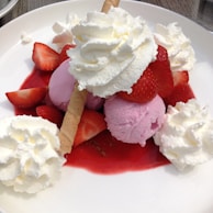 Scoops of pink ice cream topped with dollops of whipped cream and surrounded by fresh strawberries. A crispy wafer sticks out from one of the scoops. Strawberry syrup is poured over the dessert, pooling around the ice cream and fruit.