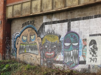 A mural with several colorful, exaggerated caricatures on a weathered brick wall. One figure has the word 'ANGRY' written above with bared teeth and furrowed eyebrows. The second face has a dark complexion with clenched teeth and yellow hair. The third caricature has a blue face with large, white eyes and sharp, exaggerated teeth. The mural also includes a small, black and white character on the far right side with the number '747' written beneath it.