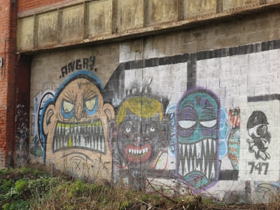 A mural with several colorful, exaggerated caricatures on a weathered brick wall. One figure has the word 'ANGRY' written above with bared teeth and furrowed eyebrows. The second face has a dark complexion with clenched teeth and yellow hair. The third caricature has a blue face with large, white eyes and sharp, exaggerated teeth. The mural also includes a small, black and white character on the far right side with the number '747' written beneath it.