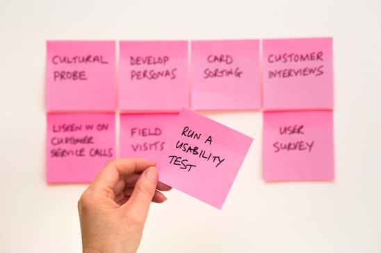 A set of pink sticky notes arranged on a wall with various handwritten tasks related to usability and customer interaction. A hand is holding one sticky note in the foreground with the writing 'RUN A USABILITY TEST'.