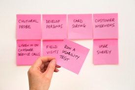 A set of pink sticky notes arranged on a wall with various handwritten tasks related to usability and customer interaction. A hand is holding one sticky note in the foreground with the writing 'RUN A USABILITY TEST'.