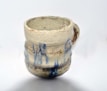 A handmade ceramic mug with a rustic appearance. The surface features a rough texture with earthy tones and blue glaze drips. The handle is small and appears to be formed organically, indicating a handcrafted design.