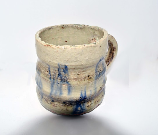 A handmade ceramic mug with a rustic appearance. The surface features a rough texture with earthy tones and blue glaze drips. The handle is small and appears to be formed organically, indicating a handcrafted design.