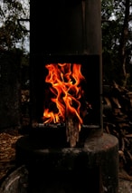 A metal stove with a black exterior contains bright, orange flames burning against a dim interior. The surrounding area includes darkened wood and metal components with trees visible in the background.