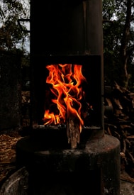 A metal stove with a black exterior contains bright, orange flames burning against a dim interior. The surrounding area includes darkened wood and metal components with trees visible in the background.